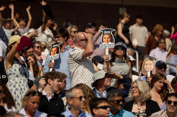 A large, animated crowd is gathered outdoors, many wearing hats and sunglasses. Some individuals are holding up signs with photos and the text "2026" and "LOOK WHO'S GRADUATING." The atmosphere appears lively and sunny.