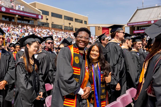 Graduates in caps and gowns celebrate at a Boston College commencement ceremony. Two individuals in the foreground smile, surrounded by a crowd of fellow graduates.