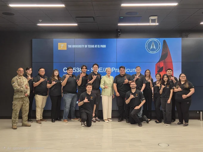 A group of people posing together in front of a screen displaying "The University of Texas at El Paso" and a rocket graphic. One individual is in a military uniform, and the others are in casual or business attire. They are making a hand gesture with their thumb and pinky finger extended. The environment appears to be a modern, well-lit room.