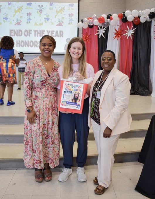 Three smiling women pose together inside a school cafeteria or multipurpose room during an end-of-year honors event. The woman in the center, Mrs. Jessica Turner, holds a colorful Speech Pathologist Day gift bag featuring a personalized appreciation graphic with her photo and name. She is wearing a light pink shirt, navy pants, and white sneakers. The woman on the left wears a long floral dress with wedge sandals and a smartwatch, while the woman on the right wears a light cream pantsuit with a colorful blouse and sandals. Behind them is a festive red, black, white, and silver balloon backdrop with starburst decorations. A projector screen in the background displays “End of the Year Honors” with Disney character graphics. Other students and staff members can be seen in the background during the celebration event.