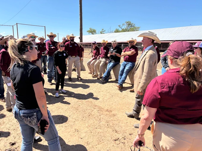 A group of people, wearing cowboy hats and casual attire, stand outside on a sunny day at a ranch or farm. They appear engaged in conversation, with several people listening to someone speaking. The background features a barn and clear blue sky.