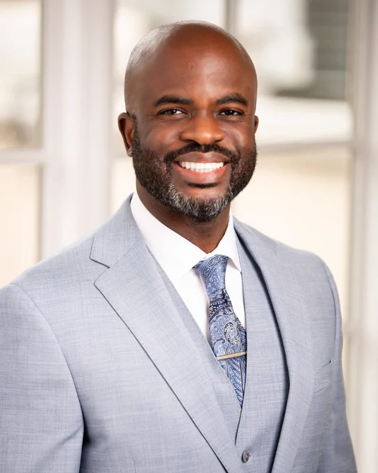 Dr. Jason A. Boothe of Tryon Medical Partners smiles wearing a suit and tie posing in a professional portrait in front of a paned glass window