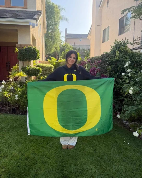 A person stands in a garden holding a University of Oregon flag.