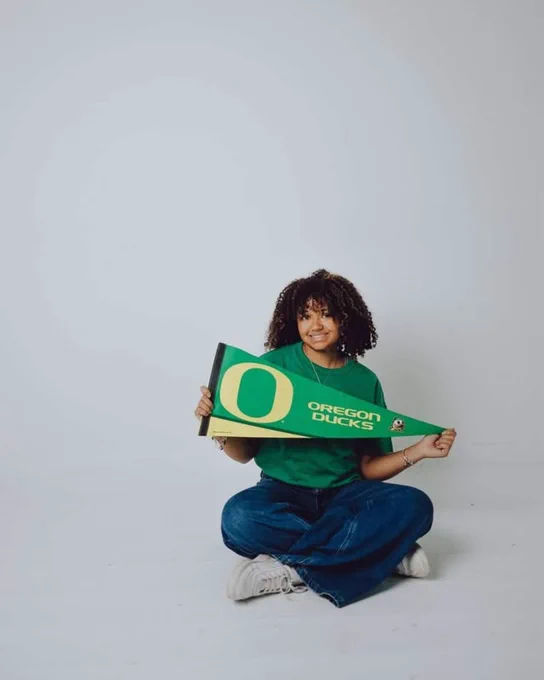 A person sitting on the floor, smiling while holding a green Oregon Ducks pennant. They are wearing a green shirt and blue jeans, with a plain white background.