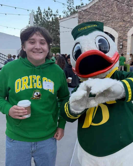 A person in an "Oregon" sweatshirt stands beside the Oregon Duck mascot. They are holding a cup and smiling. Outdoor setting with people in the background.