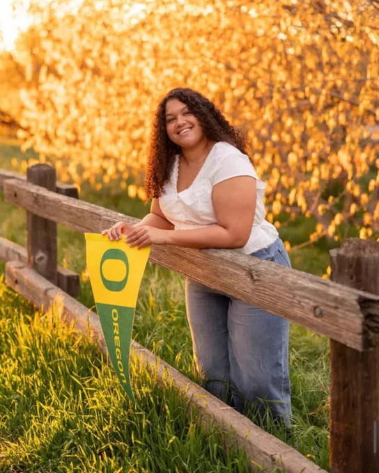 A person is leaning against a wooden fence in a grassy area with trees in the background, holding a University of Oregon pennant. The sunlight gives the scene a warm, golden glow.