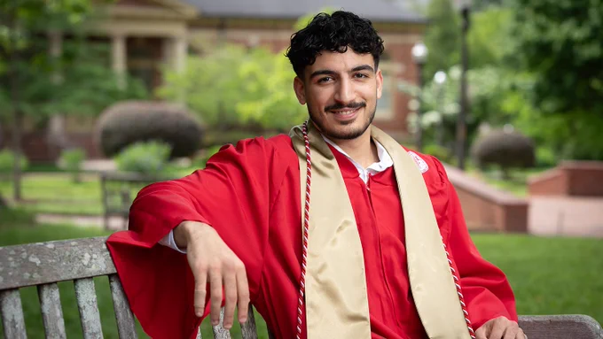 Young man in a red graduation gown and gold stole sitting on a park bench with greenery and campus buildings in the background.