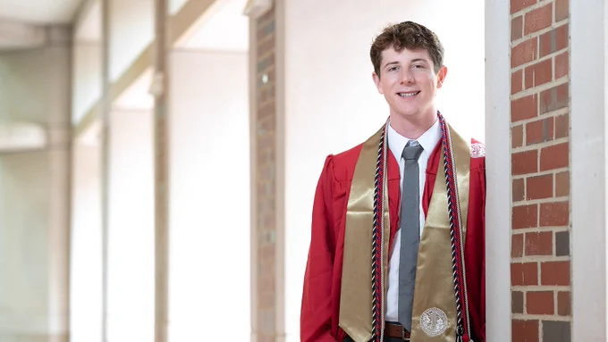 Graduate in red cap and gown with gold stole and honor cords standing against a brick wall indoors.