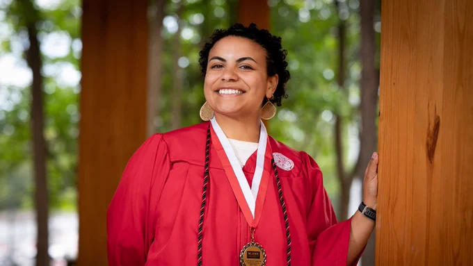 Graduate in a red cap and gown wearing a medal and honor cords, standing outdoors by wooden pillars with trees in the background.