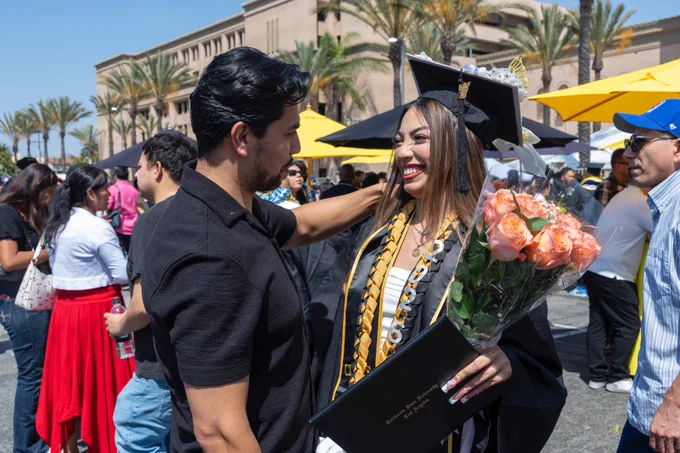 A graduate in cap and gown holds a diploma and a bouquet of orange roses, smiling while embraced by someone amidst a crowd. Palm trees and a large building are in the background, with bright yellow umbrellas.