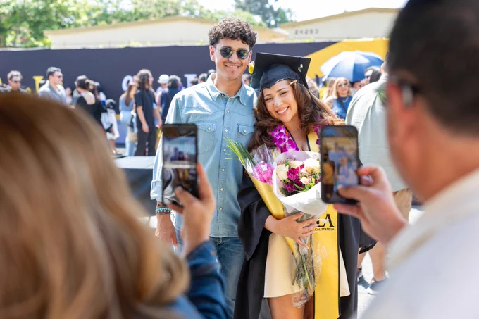 A person in a graduation cap and gown holds a bouquet of flowers, standing next to another person wearing sunglasses. They are posing for photos outdoors. In the background, partially visible, is a "CSULB" banner, indicating the location might be California State University, Long Beach.