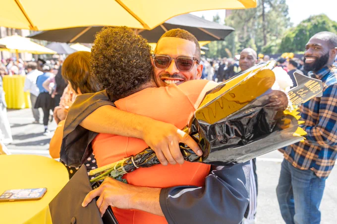 A person in a graduation gown and sunglasses joyfully hugs another person outdoors at a graduation event. They hold a bouquet and a decorated graduation cap. Other attendees are visible in the sunny background.