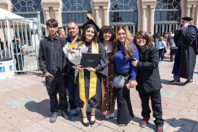 A group of people standing together outside a building, celebrating a graduation. The graduate is dressed in a cap and gown, holding a bouquet and a diploma. Other people are gathered around, smiling. The background features large windows and architectural details.