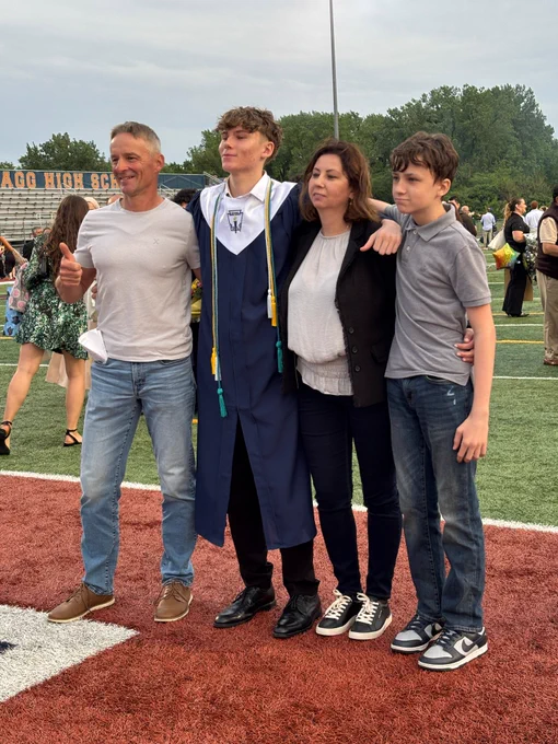 A graduate in a cap and gown stands with his family on the football field after graduation.