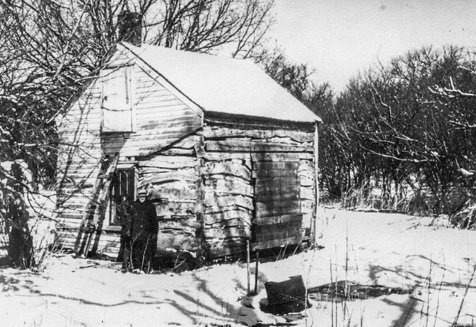 Black and white photo of a one-room log cabin. An elderly man stands in front.