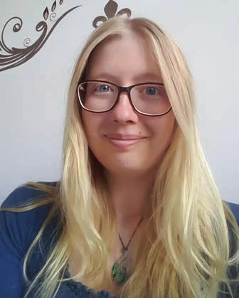 Laura Niermann pictured with long hair, wearing a blue top and a green pendant necklace against a light gray wall with decorative brown floral accents.