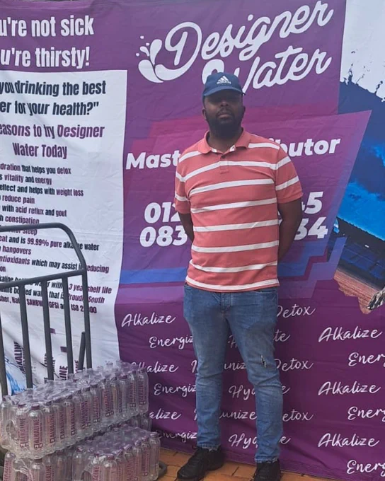 Mzimasi standing alongside a Designer Water pH10 banner and some cases of water in Centurion.