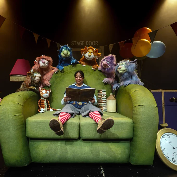 Woman sat on a large green sofa reading a book with 7 puppets listening.