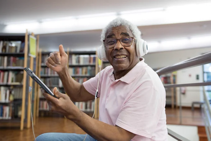 Older man in a library smiling with headphones and tablet, embracing digital learning.