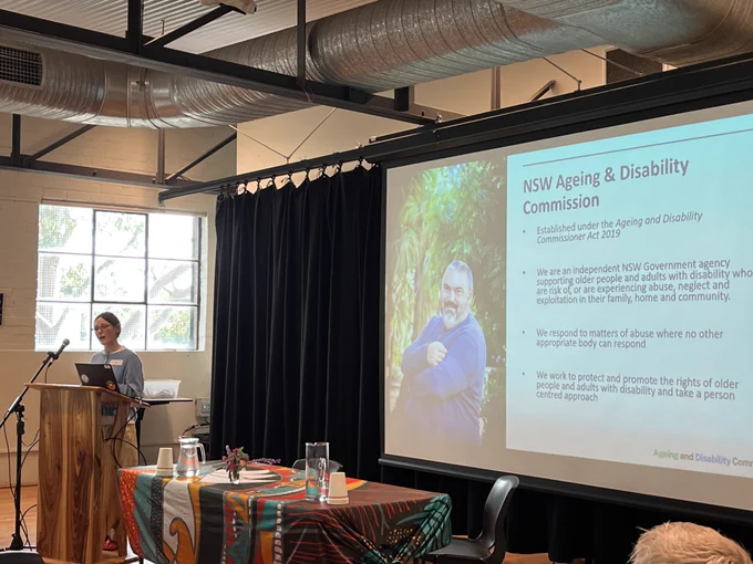 Presenter standing at a wooden lectern with a laptop and microphone, speaking to an audience. A projector screen displays “NSW Ageing & Disability Commission” with text and an image. Table with water jugs and cups in foreground.