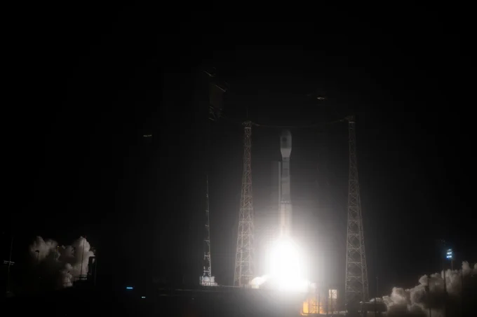 A tall, slim rocket is lifting off from a launchpad. Bright light bursts from the bottom of the rocket, and smoke billows all around the lower half of the frame. 