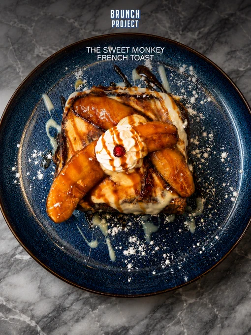 A luxurious dessert-style French toast called The Sweet Monkey French Toast served at The Brunch Project café in Koramangala, Bangalore. The image features a thick slice of golden toasted bread topped with caramelised bananas, silky whipped cream, salted caramel drizzle, chocolate sauce, and powdered sugar, plated beautifully on a textured deep blue ceramic plate placed over a marble tabletop. One creative collage layout highlights the core ingredients individually, including fresh banana slices, glossy salted caramel, soft whipped cream, and fluffy bread textures surrounding the plated French toast in the center. The second visual showcases the complete dessert in a dramatic overhead composition with rich lighting and glossy caramel textures creating a premium brunch café aesthetic. The overall mood feels indulgent, cozy, comforting, and luxurious, representing a signature sweet brunch dish from The Brunch Project known for aesthetic brunch plates, specialty coffee, and comforting caf