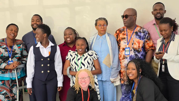 A group of delegates and disability rights advocates pose together during the 87th Ordinary Session of the African Commission on Human and Peoples’ Rights (ACHPR). The group includes men and women standing and seated indoors, with some participants using mobility aids such as crutches. Several delegates are wearing conference badges and smiling during the group photo.
