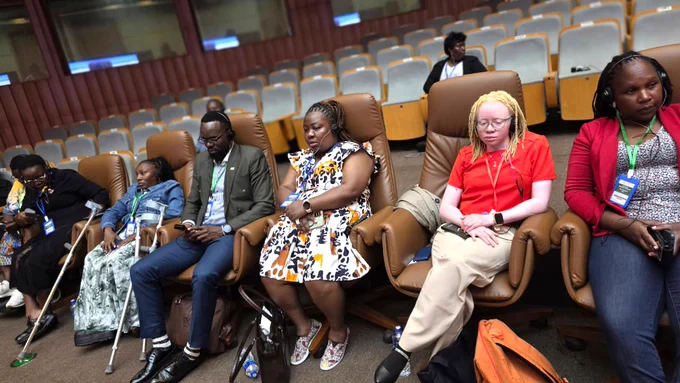 Delegates attending the 87th Ordinary Session of the African Commission on Human and Peoples’ Rights (ACHPR) sit in a conference hall wearing headsets during a session. The group includes men and women with disabilities seated in accessible chairs, with some participants using mobility aids such as crutches. Rows of seats are visible in the background of the meeting venue.