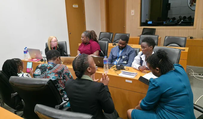 Delegates from disability rights organisations engage in a discussion around a conference table during a meeting session at the 87th Ordinary Session of the African Commission on Human and Peoples’ Rights (ACHPR). Participants are seated with laptops, notebooks, and headsets as they collaborate in a meeting room setting.