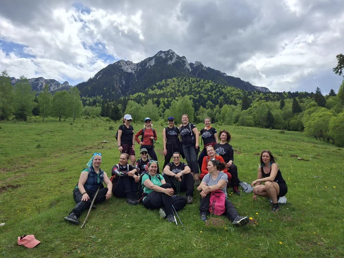 A group of women hikers resting in a green mountain meadow beneath dramatic cloudy skies and forest-covered peaks in Romania. The group is smiling and gathered together after a hike, surrounded by spring vegetation and alpine scenery, reflecting friendship, community, and shared outdoor experiences.