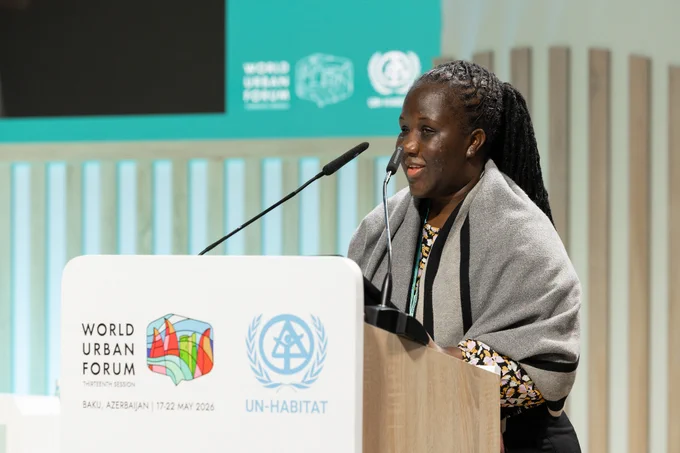 A woman speaks at a podium during the World Urban Forum Thirteenth Session in Baku, Azerbaijan. She is addressing the audience with microphones in front of her, wearing a patterned outfit and a gray shawl. The podium displays the World Urban Forum and UN-Habitat logos, with event branding visible on the screen behind her.