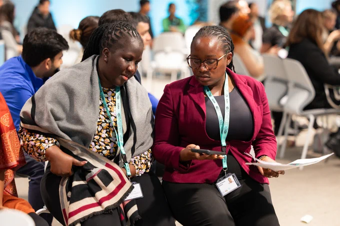 Two women wearing conference badges sit together in a discussion area at an event. One woman, wrapped in a gray shawl, leans in while the other, wearing glasses and a burgundy blazer, holds a phone and papers as they review information together. Other attendees are visible in the background in a busy conference setting.