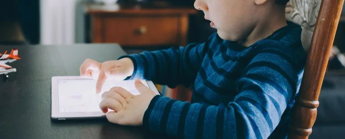 A young boy sitting on a wooden chair at a dining room table using a tablet.