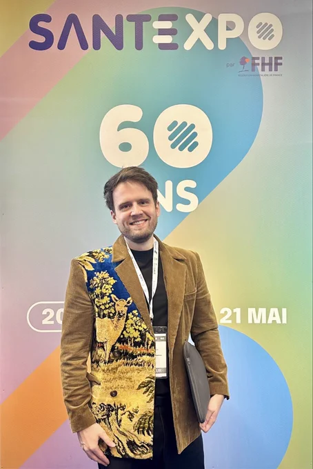 Portrait of Benjamin André-Micolon at Santexpo 2026 in Paris. He stands smiling in front of the event signage, wearing a brown jacket with an illustrated fabric panel and a conference badge, carrying a laptop sleeve while attending meetings at the healthcare technology event.