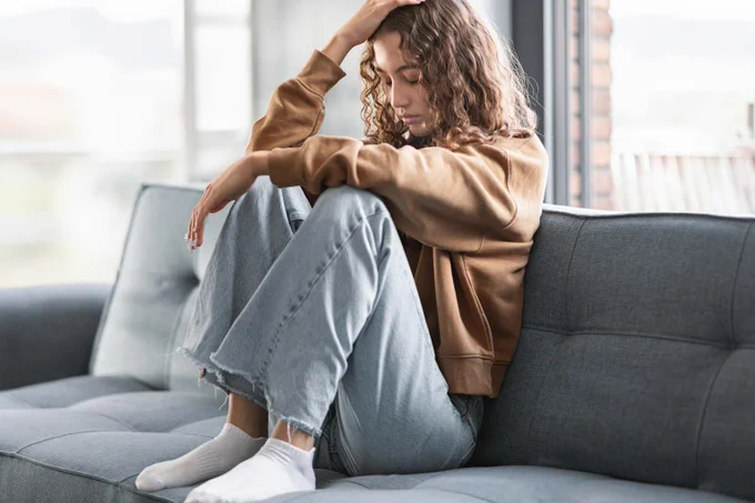 A person with curly hair sits on a gray sofa, wearing a brown jacket and light blue jeans. The individual appears to be in a contemplative mood, gazing downward in a softly lit room.