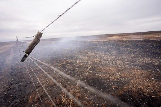 A burnt field stretches into the distance under a cloudy sky. A barbed wire fence runs diagonally across the foreground. Smoke lingers above the landscape.