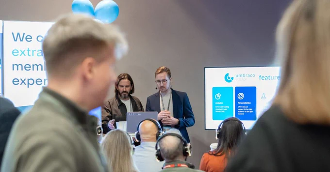 Two presenters stand behind a laptop at a tech event, while an audience wearing headphones listens in the foreground.