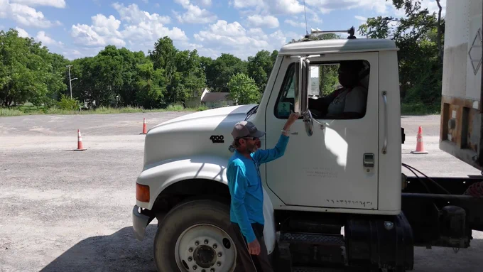 Man in blue shirt reaching up to touch the door handle of a white semi-truck parked on a sunny day.