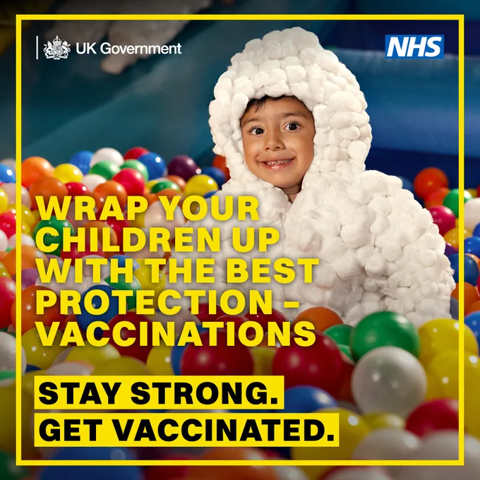 Child dressed in a white protective suit sitting in a colorful ball pit, promoting vaccination for children by UK Government and NHS.