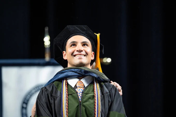 A person wearing a graduation cap and gown smiles while looking upward. A stole, cords, and tassel are part of the graduation attire. In the background, there is a blurred banner with an indiscernible emblem.