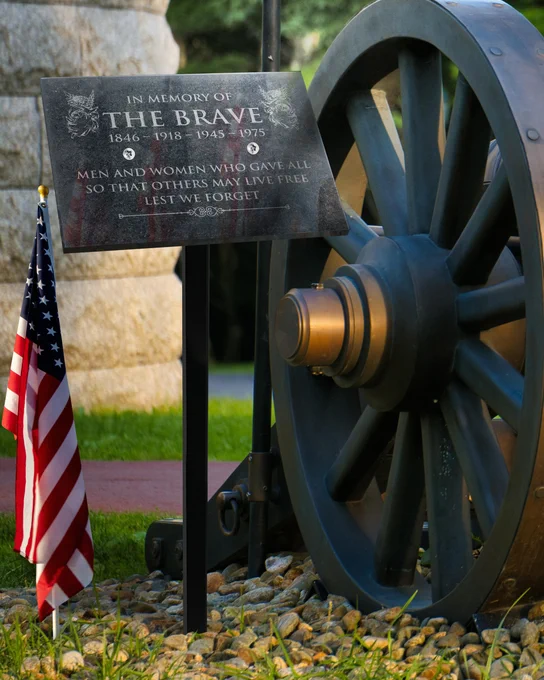 Granite Plaque Engraved With Memorial Details Displayed Outdoors Via Sign Stand Beside American Flag And Vintage Cannon