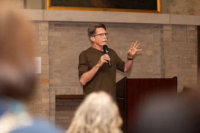 Rick Bayless holding a microphone and gesturing during a presentation in the Jane Addams Hull House