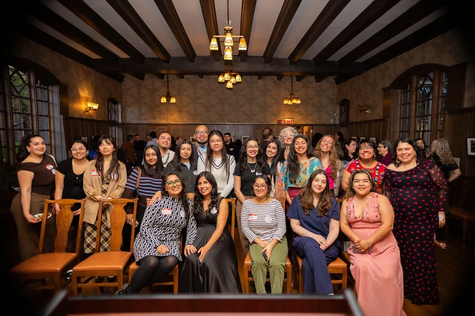 Group photo of diverse women and men smiling in a warmly lit room with wooden beams and vintage chandeliers.