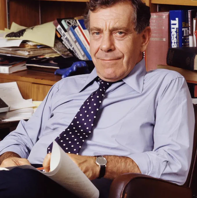 Morley Safer sits in a leather chair in an office, holding a stack of papers. He wears a light blue dress shirt with a dark polka dot tie and a wristwatch. Behind him, a wooden desk and shelves are filled with books, folders, and loose papers. A typewriter sits on the desk to his left, and several books are stacked on the shelves, including large hardcover volumes.

