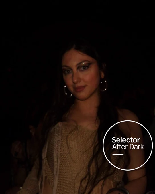 Portrait of LILITH wearing hoop earrings and a knit top at a dimly lit nighttime event. The Selector After Dark logo sits in a white circle at the bottom right.