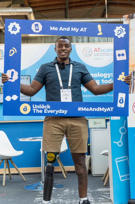 A man with a prosthetic leg stands smiling inside a large blue “Me And My AT” photo frame at an exhibition booth. He is wearing a dark shirt, shorts, and an event badge, with ATscale branding visible in the background. The frame features icons representing different types of assistive technology and includes the text “Unlock The Everyday” and “#MeAndMyAT.