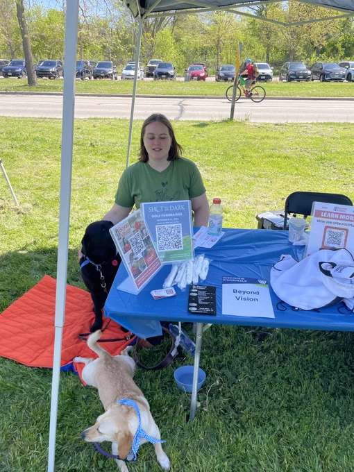 Rose at the Beyond Vision booth with her dog Terra laying out in front on the grass.