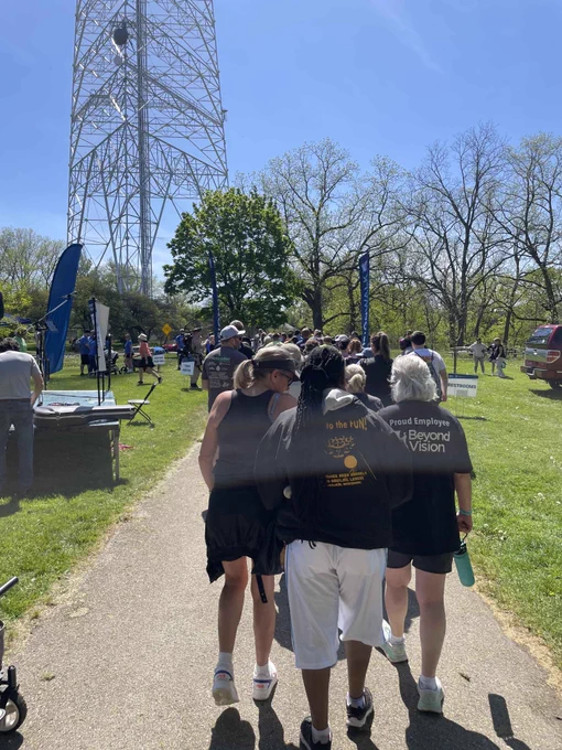 Group of people walking along a park path on a sunny day near a tall metal tower and green trees.