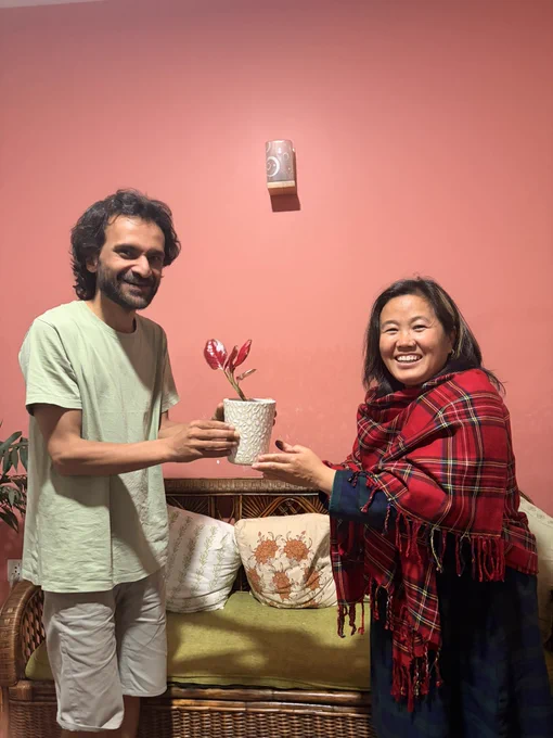 Two people are exchanging a white flower pot with red flower plant. They both smile for the picture while they are standing. The background is a dark pink wall.