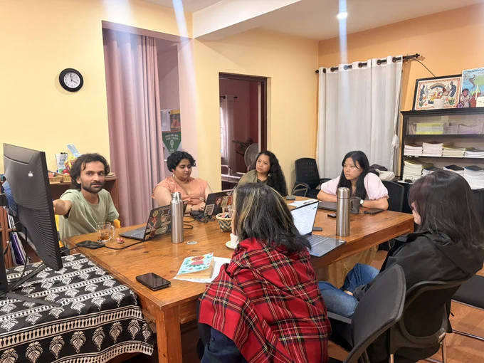 A zoomed out click of the room where six people are sitting around a table while looking at the screen. The background is the office space of Body & Data which has yellow walls.